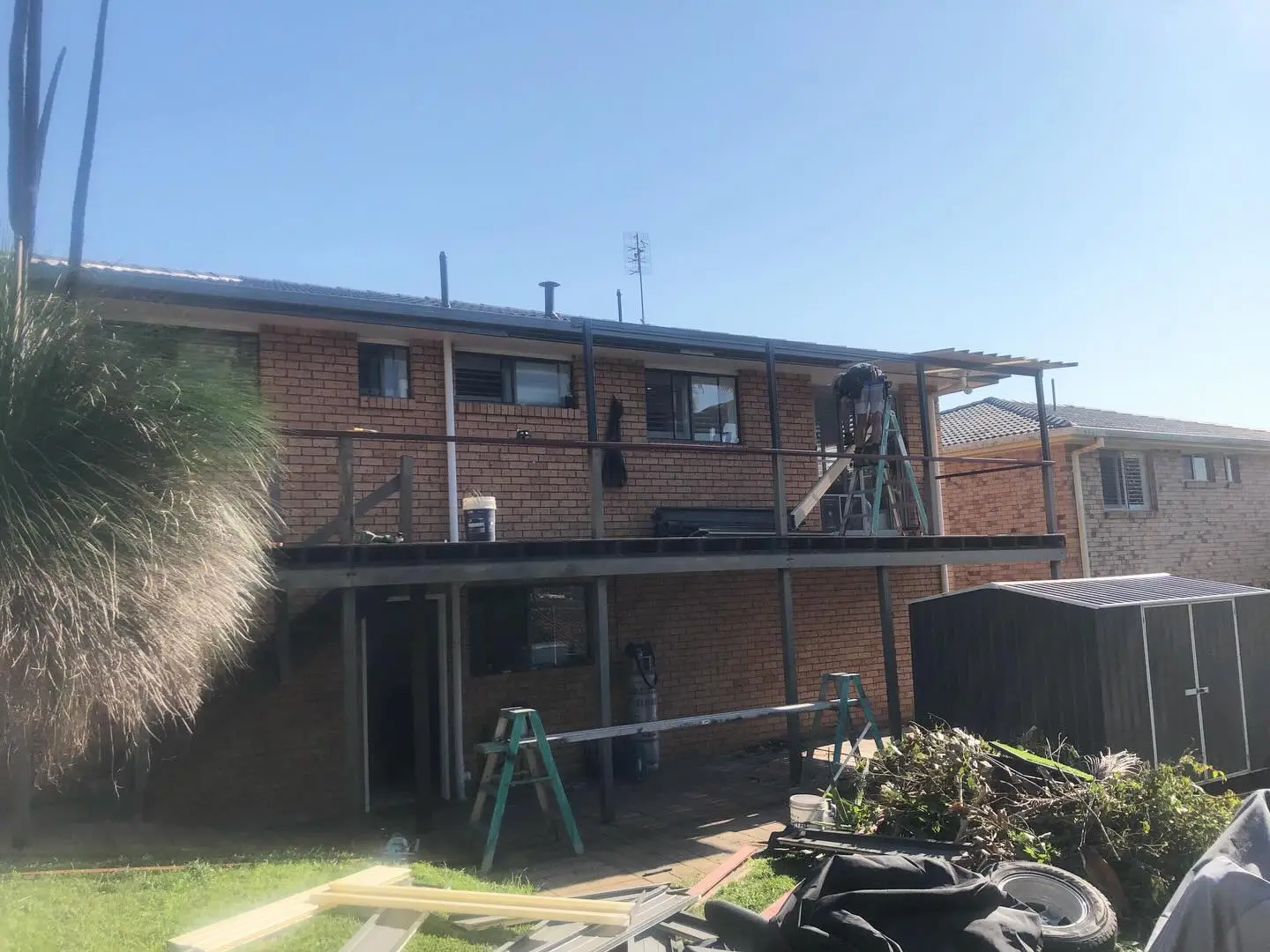 The back of a two-storey brick house before the job: the old balcony and pergola being stripped by a worker on a ladder, a garden shed, and green waste and sheet offcuts piled on the lawn.