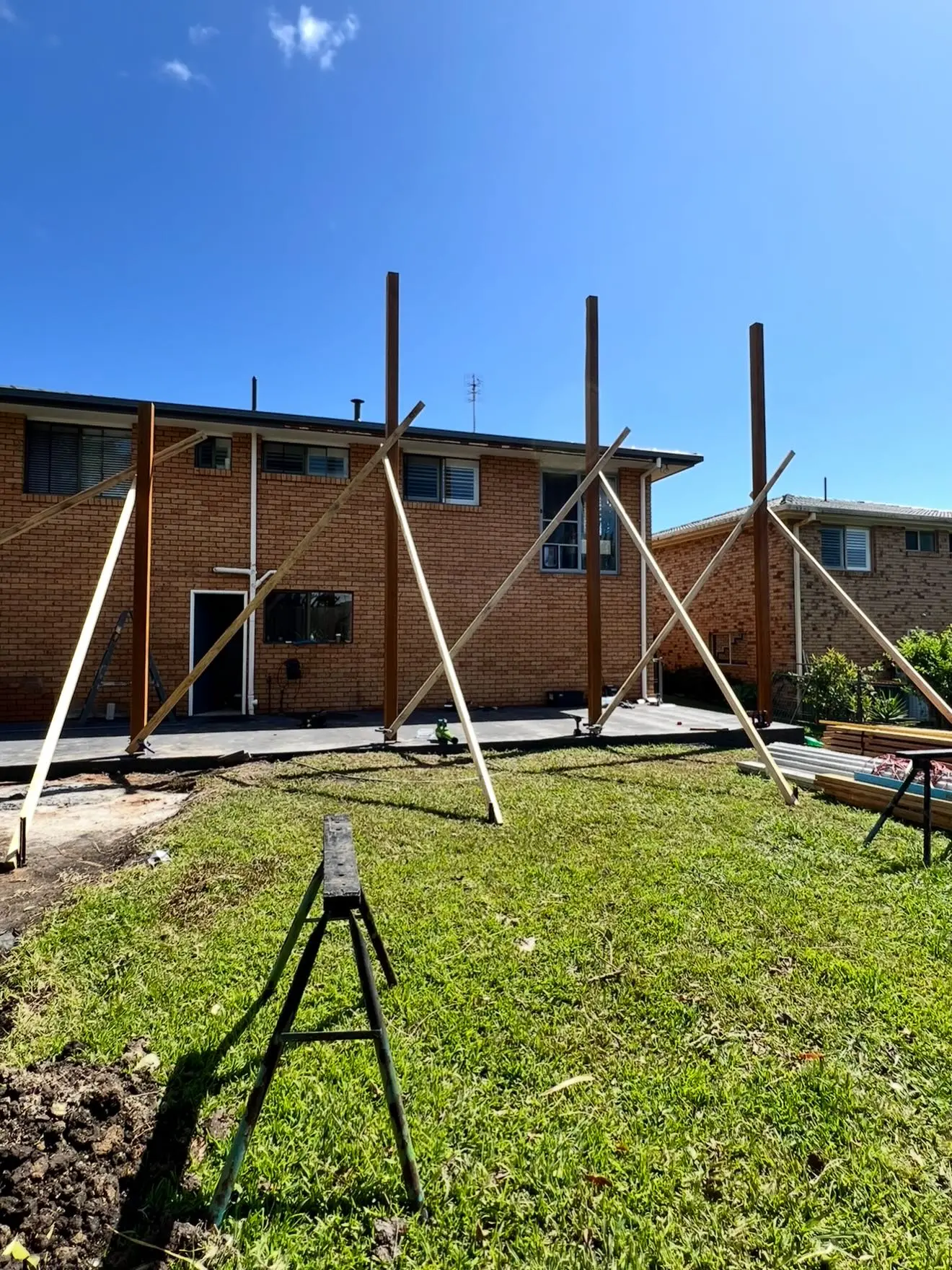 Four tall timber posts standing on a new slab at the back of the brick house, each braced with crossed props, under a deep blue sky.