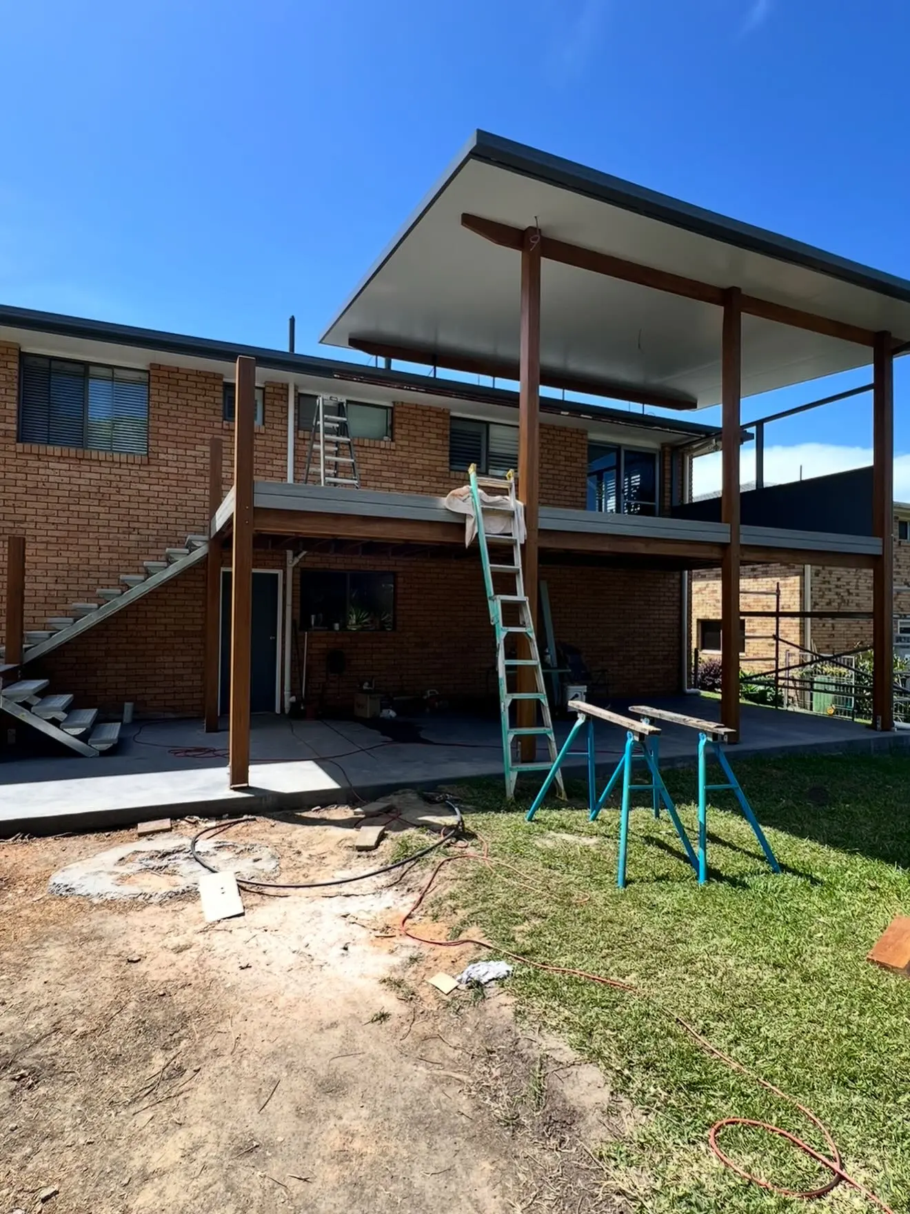 The back of the house with the deck boards down and a flat roof up on timber posts and beams, a new stair at the left, bare dirt and patchy lawn in front.
