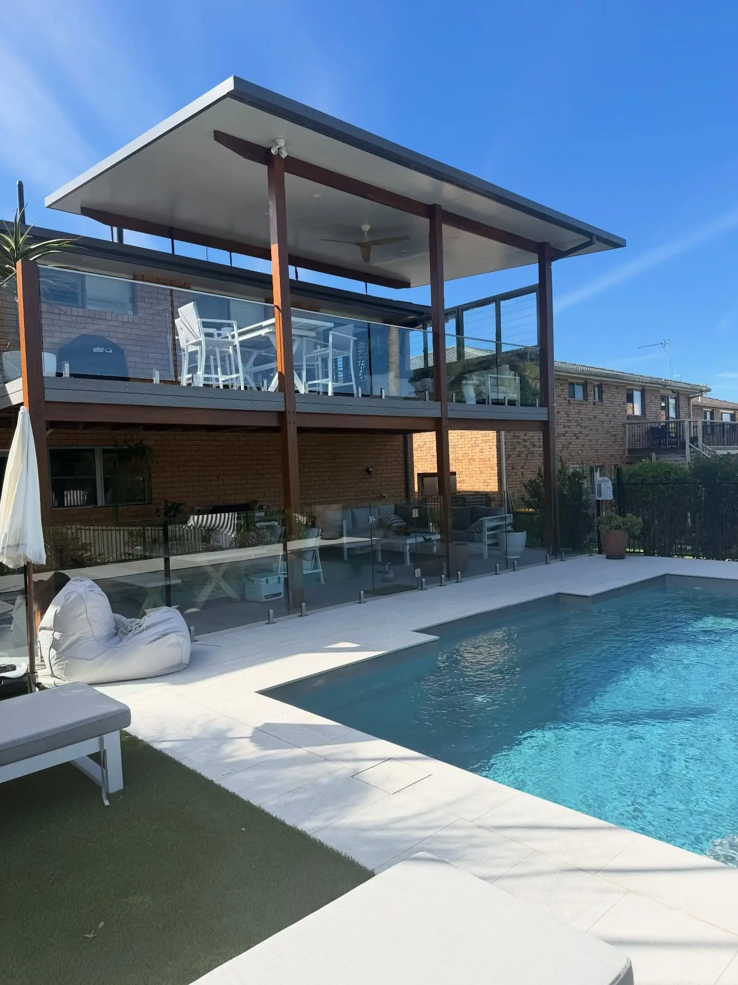 The finished backyard: an elevated deck under a flat roof on tall timber posts, glass balustrade, a white outdoor setting on the deck, a lounge underneath, a glass pool fence and pale paving around a blue pool.