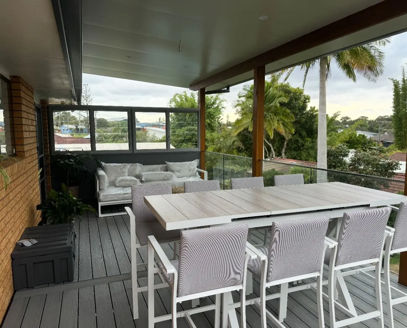 On the finished deck: a bar-height outdoor table and chairs on grey boards, a lounge still in its wrapping, glass balustrade, a timber post and beam, palms and rooftops beyond.