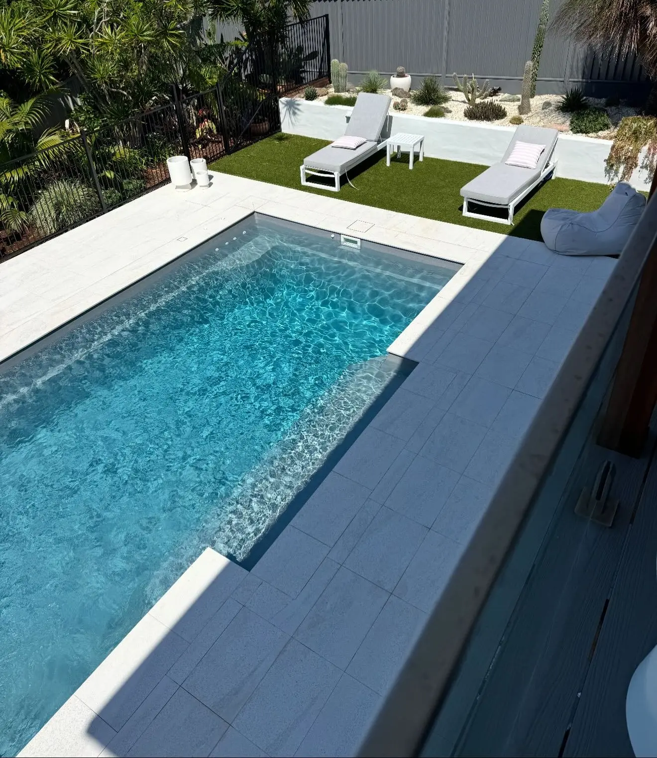 Looking down from the deck at a rectangular pool with rippling water, pale coping and paving, a strip of turf with two sun loungers and a white planter of cacti against a grey fence.