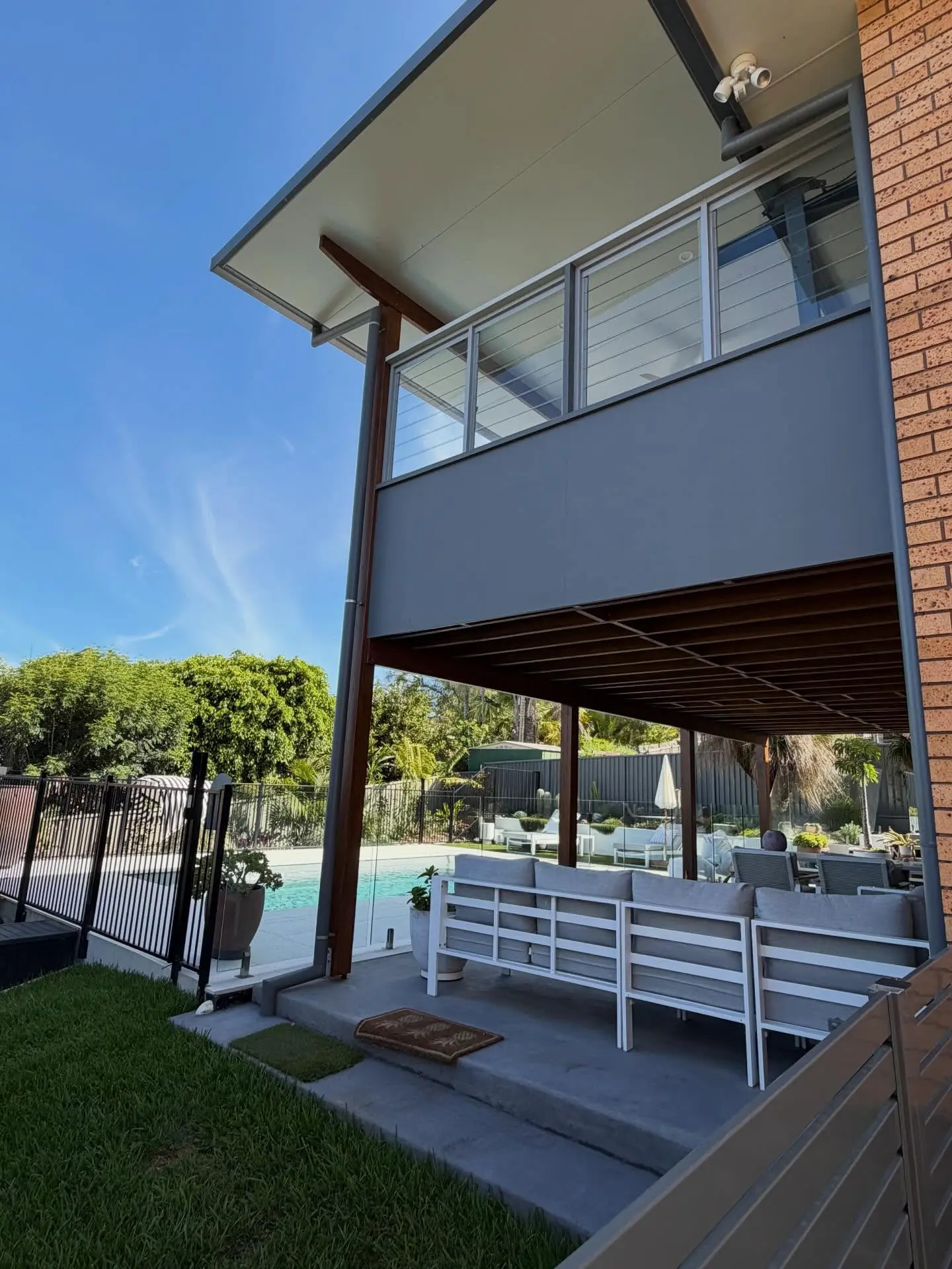 The finished deck from the side: the flat roof oversailing on a timber post, a grey privacy wall with a wire screen above, exposed timber joists underneath, an outdoor lounge on the slab and the pool beyond.