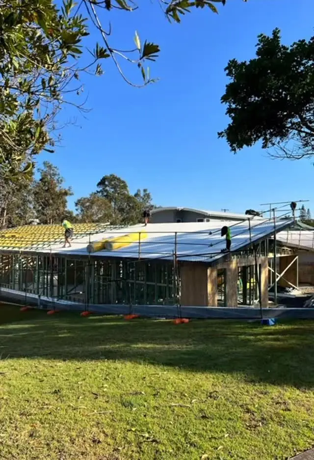 A single-storey house mid-build seen across a lawn: wall frames standing behind edge scaffold and the roof going on over yellow battens, gum trees and a clear blue sky.