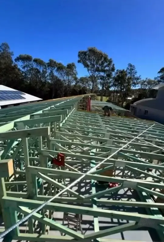 Looking along a run of roof trusses from above, a worker crouched on the far side with his back turned, gum trees and blue sky behind.