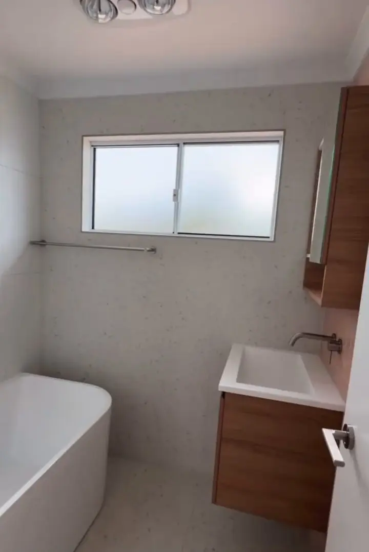 A bathroom with a freestanding bath, a wall-hung timber-look vanity with a white basin, speckled wall tiles and a frosted window.