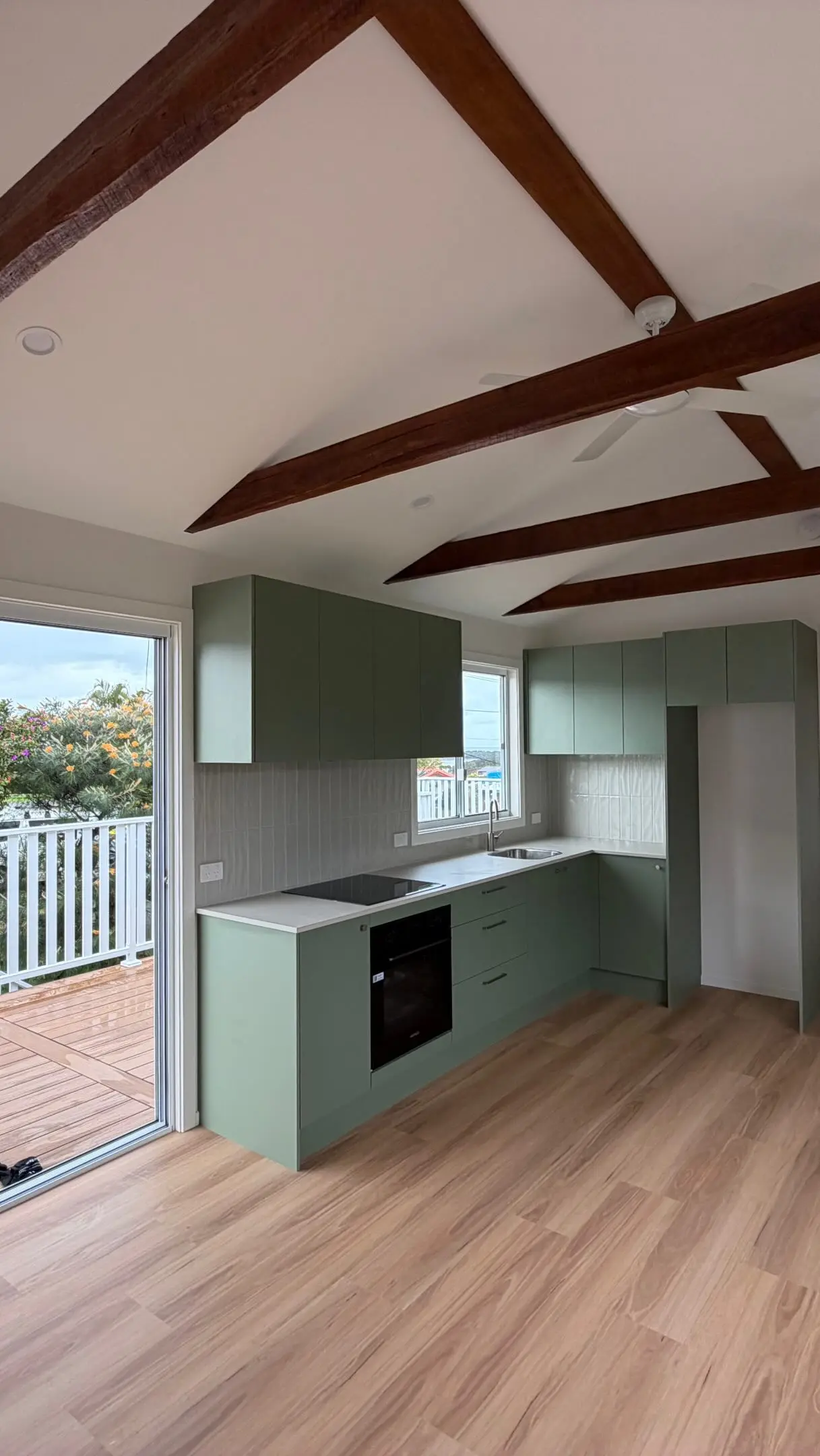 A small kitchen in sage green cabinetry with a white benchtop and pale tiled splashback, a timber-look plank floor, a white raked ceiling crossed by dark exposed rafters, and a sliding door open to a timber deck with a white balustrade.