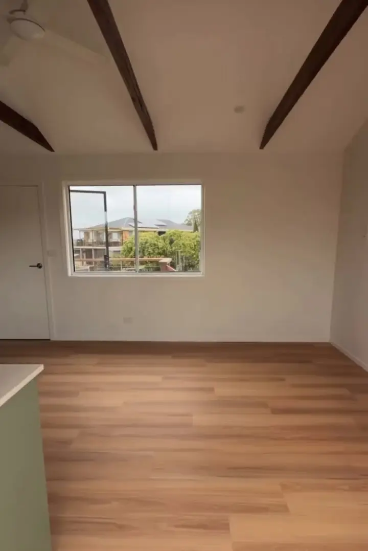 An empty room with a timber-look plank floor, white walls, a window and a white raked ceiling crossed by dark exposed rafters.