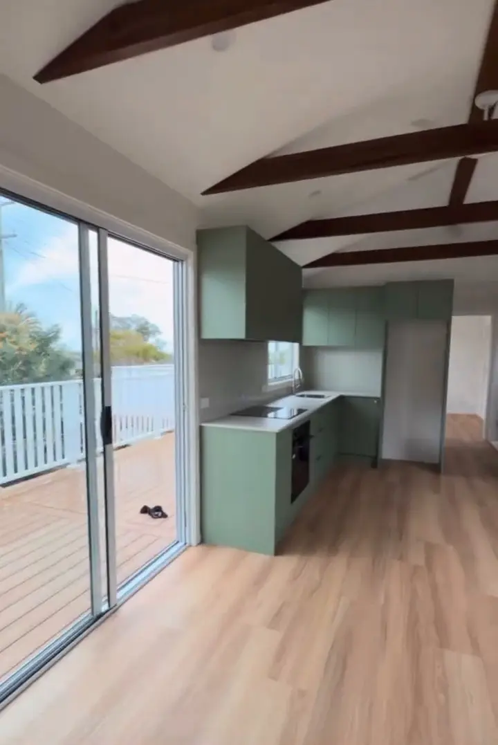 The main room from the far end: sage green kitchen cabinetry along one wall, a timber-look plank floor, a white raked ceiling crossed by dark exposed rafters, and a sliding glass door open to a timber deck with a white balustrade.