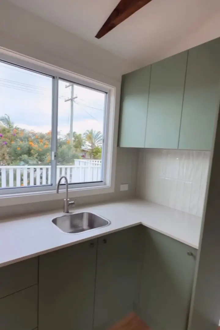 The corner of a sage green kitchen: a white benchtop with an inset sink under a window, overhead cupboards and a pale tiled splashback.