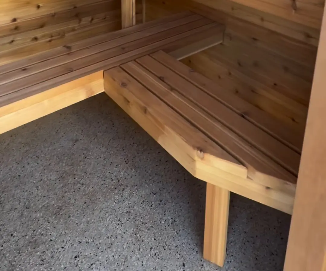 The corner of an L-shaped slatted timber bench in a timber-lined sauna, on a speckled concrete floor.