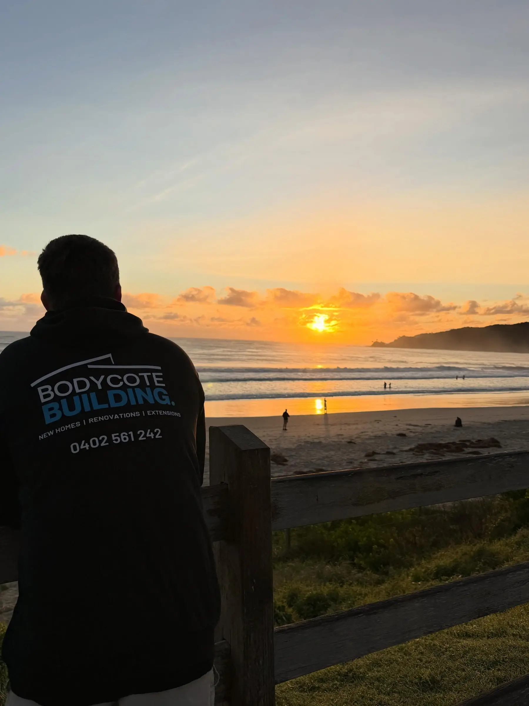 Seen from behind, a person in a Bodycote Building hoodie leans on a timber rail as the sun comes up over a beach and a headland.