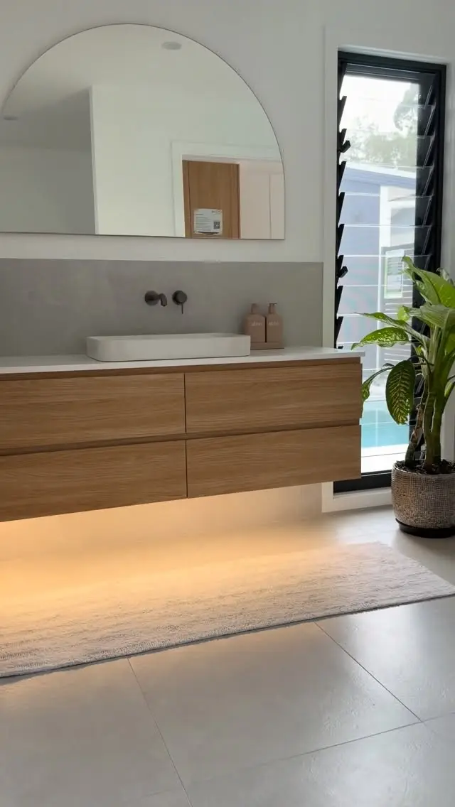 A bathroom vanity wall: floating timber-look drawers with a warm light strip underneath, a white basin, a wall mixer, a half-arch mirror and a black-framed louvre window.
