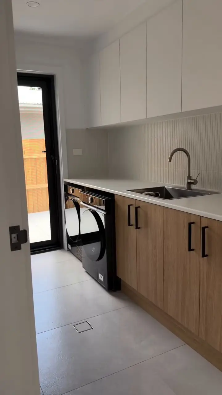 A laundry with timber-look base cupboards, a white benchtop with an inset sink, white overhead cupboards, narrow vertical tiles on the splashback, two black front-loading machines under the bench and a glazed door to outside.