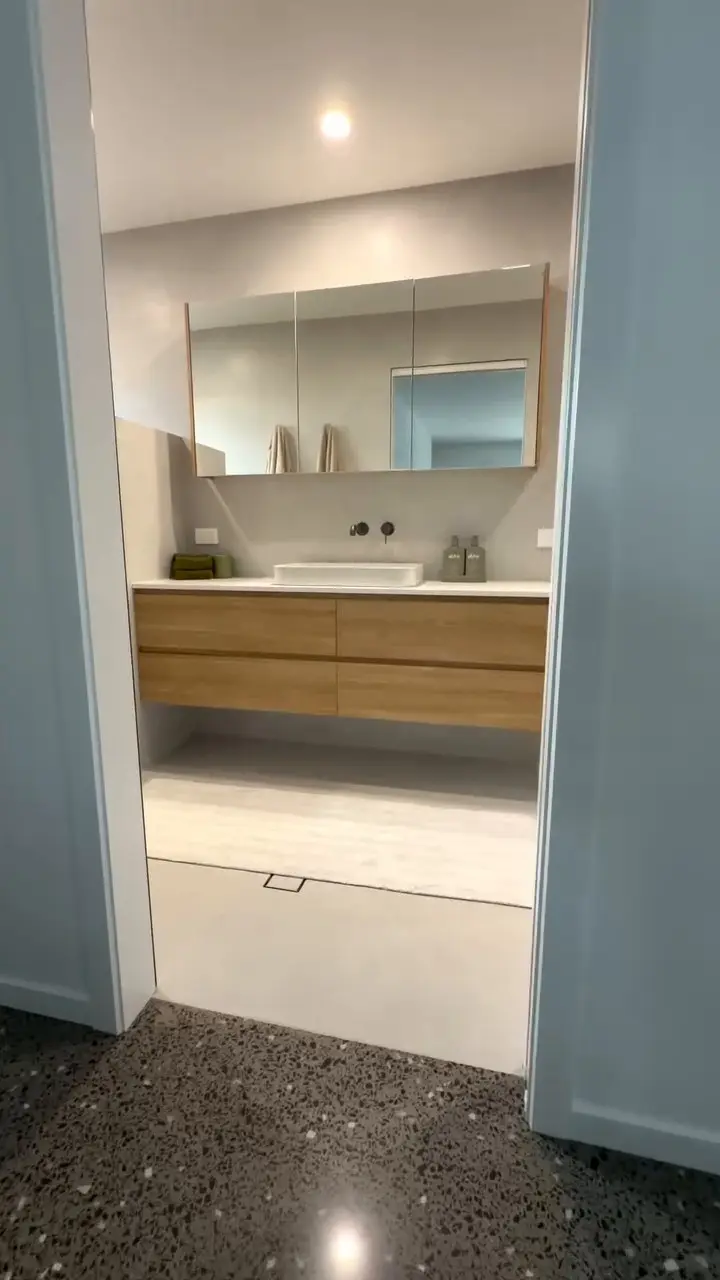 Looking through a doorway into a bathroom: dark polished concrete with pale stone chips runs flush into the floor tiles with no step, a floating timber-look vanity and a mirrored cabinet beyond.