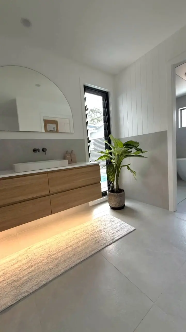A bathroom with a floating timber-look vanity lit from below, a half-arch mirror, a black-framed louvre window and a potted plant, and white VJ panelling on the wall above half-height grey tiles.